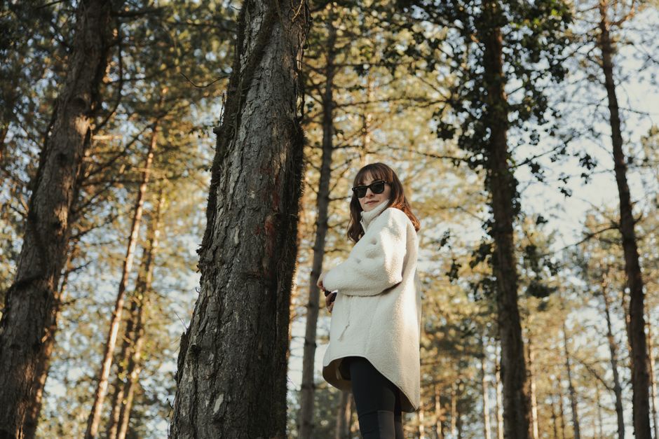 Woman in sunglasses standing confidently amidst tall trees in the forest.