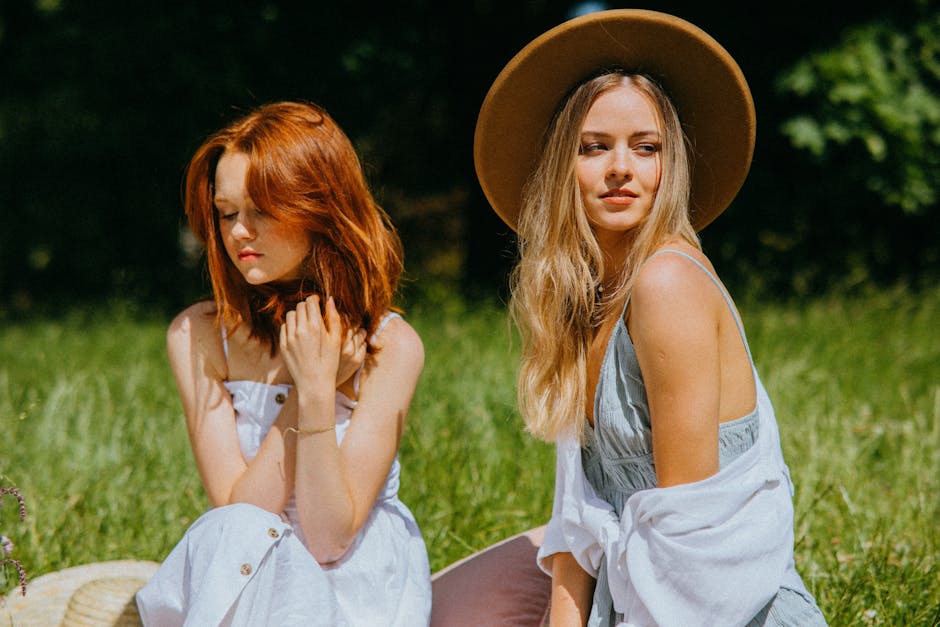 Two women in summer dresses enjoying a sunny day outdoors, sitting on grass.