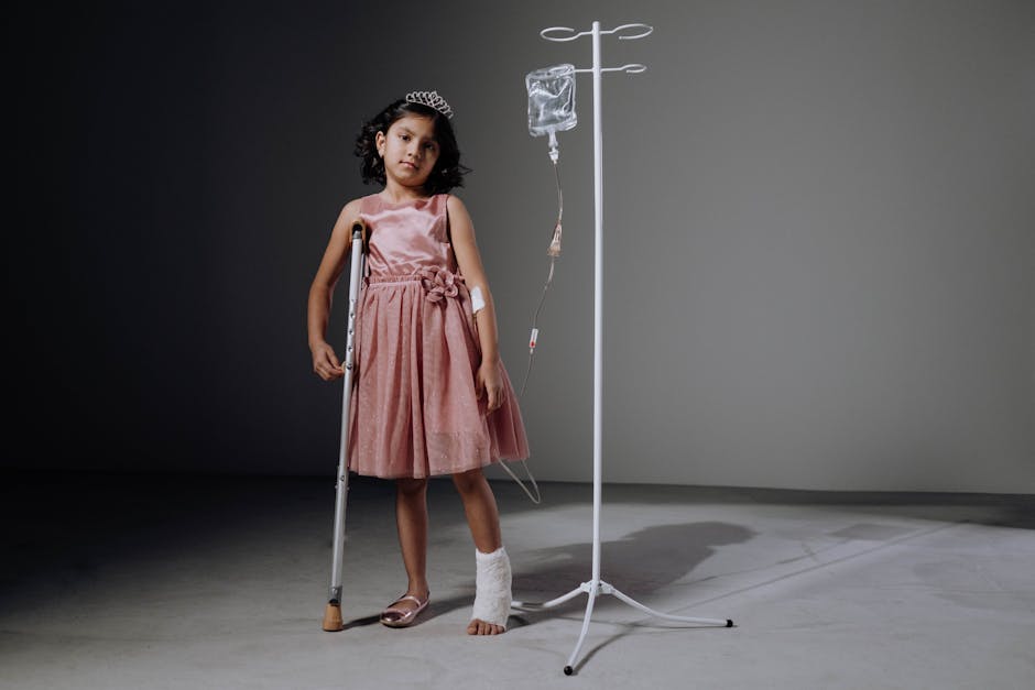 Injured girl in pink dress using crutches next to an IV stand in a studio setting.
